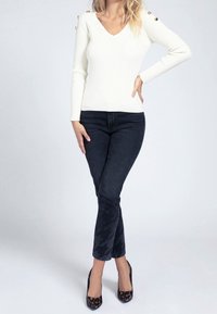 Weißer, gerippter Strickpullover mit V-Ausschnitt und goldfarbenen Knöpfen an den Schultern, kombiniert mit dunklen Jeans mit Hahnentrittmuster und bedruckten Absätzen.