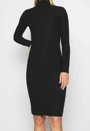 Kobieta ubrana w dopasowaną czarną sukienkę midi z długim rękawem i golfem, stojąca z jedną ręką na biodrze na jednolitym jasnym tle.