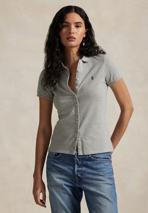 Femme aux longs cheveux bouclés portant un polo gris ajusté à boutons et un jean bleu, debout devant un fond beige uni.