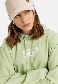 Felpa verde menta con coulisse, caratterizzata da un logo a cuore bianco e oro, indossata con un cappellino beige. Tessuto liscio e vestibilità comoda.