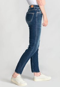 Jeans de mezclilla en azul oscuro con un corte ajustado. Las características incluyen cinco bolsillos, costuras en contraste y una etiqueta de cuero marrón en la cintura.
