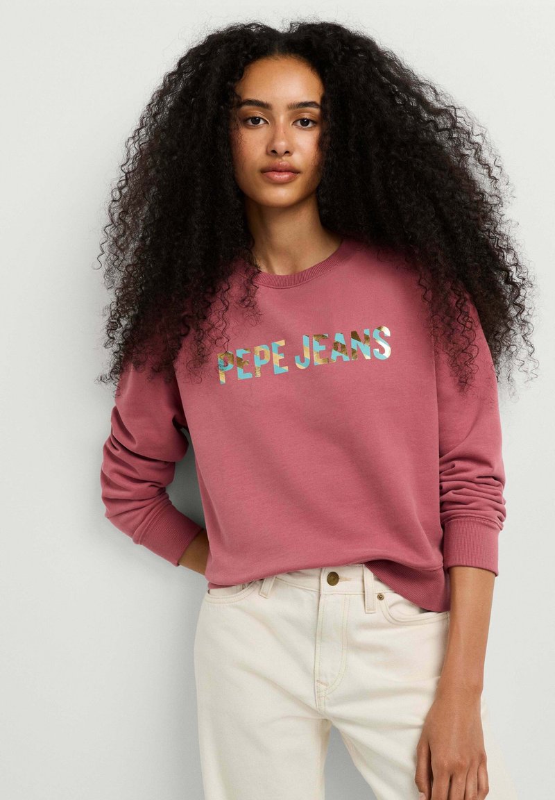 Joven mujer con cabello largo y rizado, vestida con una sudadera rosa de "PEPE JEANS" y pantalones de color claro, de pie frente a un fondo sencillo.