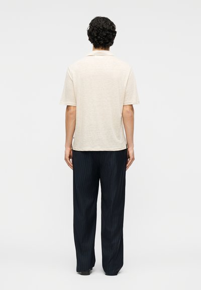 Homme aux cheveux bouclés et foncés, portant un polo crème à manches courtes et un pantalon ample à rayures fines foncées, debout de dos.