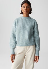 Femme aux cheveux courts portant un pull en maille bleu clair et un pantalon crème, se tenant devant un fond léger uniforme.