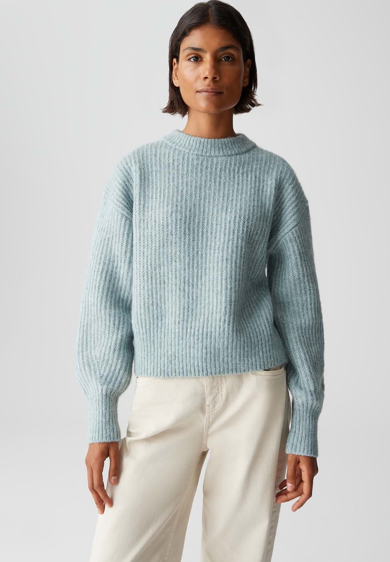 Femme aux cheveux courts portant un pull en maille bleu clair et un pantalon crème, se tenant devant un fond léger uniforme.