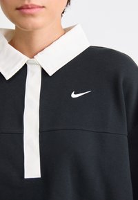 Čierna mikina s bielym golierom a prednou légou. Obsahuje malé biele logo Nike na ľavej hrudi. Textúrovaný materiál.