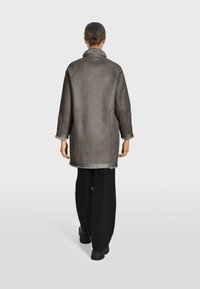 Sivi kaput napravljen od mekog, teksturiranog materijala s širokim kragnom, spuštenim ramenima i bočnim džepovima, uparen s crnim hlačama širokih nogavica.