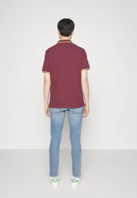 Mörkröd polo skjorta med guldsnor, parat med ljusblå jeans och vita skor med gröna detaljer; enkel, klassisk avslappnad stil.