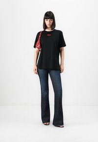 Woman with straight dark hair wears black oversized t-shirt, dark flared jeans, black sandals, and carries small red shoulder bag.