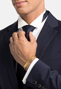 Pulsera de oro con un charm rectangular plano y una cadena enlazada, llevada sobre un traje azul marino, con una camisa blanca y una corbata de patrones oscuros.