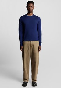 Camicia blu navy a maniche lunghe con scollo a girocollo, con un piccolo logo sul petto. Abbinata a pantaloni larghi color sabbia e calzature nere.