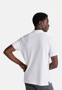 Camisa polo blanca de manga corta, con cuello de canalé y dobladillo recto. El modelo está de pie con las manos en las caderas, mostrando la vista trasera.