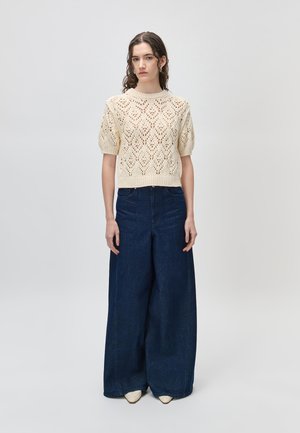 Kvinne med krøllete hår iført en beige kortermet strikket genser og høytlivede vide mørkeblå jeans som står mot en ensfarget bakgrunn.