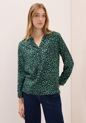 Femme avec de longs cheveux blonds portant un blouse à manches longues noire et verte à motifs et un jeans bleu foncé, se tenant contre un fond clair et uni.