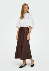 Chemise blanche oversize à manches courtes, associée à une longue jupe en cuir marron. Des escarpins noirs à talon ouvert complètent la tenue.