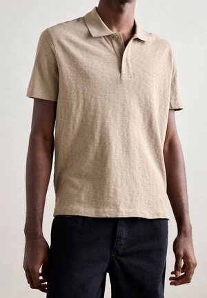 Polo beige hecho de un tejido texturizado, con cuello y mangas cortas. Marca sutil en el pecho. Combinado con jeans oscuros.