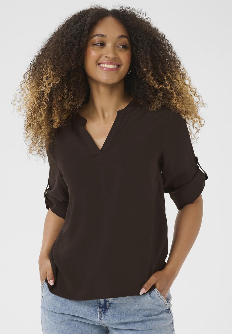 Blouse noire à manches courtes avec un col en V et des poignets roulés, offrant une texture lisse et une coupe décontractée. Le mannequin pose avec les mains dans les poches.