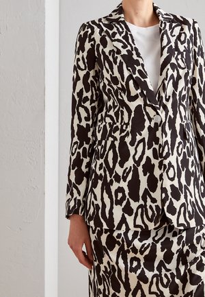 Frau trägt einen schwarz-weißen Blazer mit Tierdruck, passenden Rock und ein weißes Hemd darunter und steht vor einer weißen Wand.