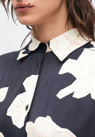 Μπλε ναυτικό πουκάμισο με μεγάλα λευκά λουλουδένια σχέδια, αντίθετο κρεμ γιακά και μαύρα κουμπιά, κατασκευασμένο από λείο και αναπνεύσιμο ύφασμα.
