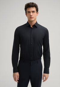 Schwarzes Langarmhemd mit glatter Textur, durchgehender Knopfleiste und spitzem Kragen, kombiniert mit dunklen Hosen.