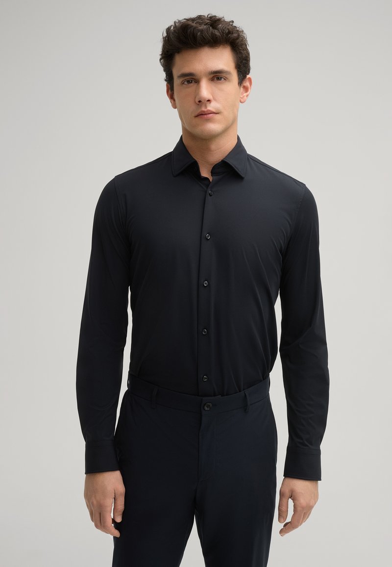 Schwarzes Langarmhemd mit glatter Textur, durchgehender Knopfleiste und spitzem Kragen, kombiniert mit dunklen Hosen.