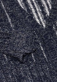 Cardigan lavorato a maglia di colore blu navy con texture a traforo caratterizzata da intricati motivi a treccia e accenti metallici intrecciati nel tessuto.