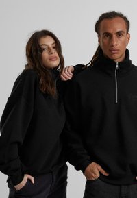 Svart hettejakke med glidelås laget av mykt materiale, med høy krage, avslappet passform og ribbede mansjetter. En liten logo er synlig på brystet.