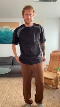 Camisa preta de mangas curtas com um bolso e detalhes cinzentos nos ombros, combinada com calças castanhas folgadas. Traje casual num chão texturizado.