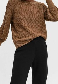 Pull à manches longues marron texturé associé à un pantalon noir taille haute porté par une personne debout sur un fond uni.