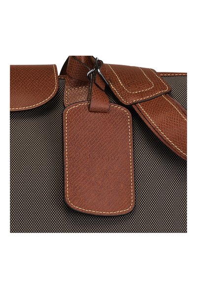 Braunes strukturiertes Lederetikett, das an einer dunklen gewebten Stofftasche befestigt ist, mit einer gesteppten Kante und eingeprägtem Logo. Metallbeschläge sichtbar.