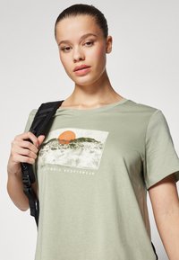 Šviesiai žalia plačiakraščio trumpomis rankovėmis marškinėliai su kalno ir saulės grafika, pasižymintys laisvu kirpimu ir minkšta tekstūra. Logotipas skelbia "Columbia Sportswear."