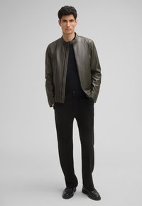 Olivenfarbene Lederjacke mit rundem Kragen, Reißverschluss und gerippter Textur, kombiniert mit schwarzen Hosen und schwarzen Slippern.