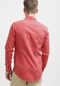 Rotes Hemd mit langen Ärmeln aus einem strukturierten Baumwollmix. Verfügt über einen spitzen Kragen und geknöpfte Manschetten. Der Saum ist leicht abgerundet.