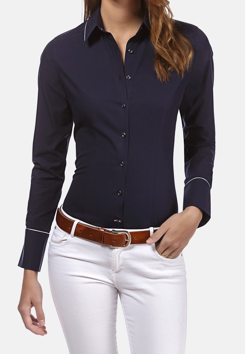 Marineblaues Hemd mit langen Ärmeln und kontrastierenden weißen Nähten an den Manschetten, kombiniert mit weißen Jeans und einem braunen Ledergürtel.