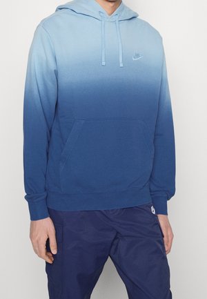 Homme portant un sweat à capuche bleu ombré avec poche avant et pantalon bleu assorti, debout devant un fond clair uni.