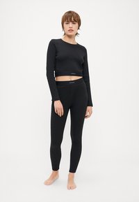 Svart kortermet topp med lange ermer og høyt liv leggings, begge laget av elastisk stoff, med en subtil logo-bånd ved midjen.