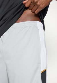 Ανοιχτό γκρι αθλητικές βερμούδες με μαύρη και λευκή πλαϊνή ταινία και χρυσή πινελιά. Διαθέτουν ελαστική ζώνη μέσης και αναπνεύσιμο υλικό.