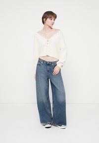Cremfarbener, kurz geschnittener Pullover mit Spitzenakzenten und Knopfverschluss, kombiniert mit weit geschnittenen, blauen Jeans. Schwarz-weiße Sneakers runden den Look ab.