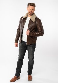 Giubbotto bomber in pelle marrone con colletto in shearling beige, polsini a costine e design aderente, indossato sopra una camicia bianca e abbinato a jeans scuri.