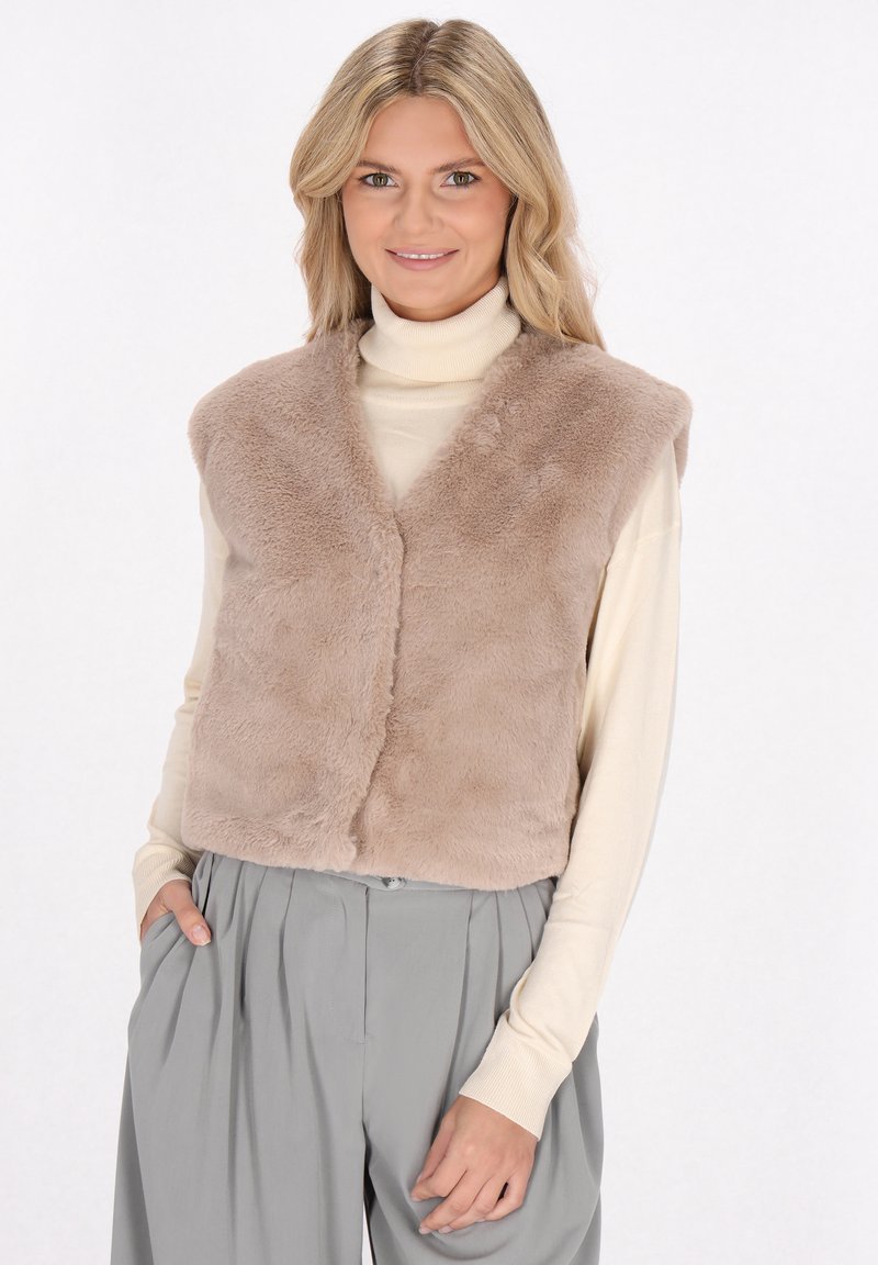 Beige Kunstfellweste mit V-Ausschnitt, ohne Ärmel, weiche Textur und frontale Verschluss. Kombiniert mit einem cremefarbenen Rollkragenpullover und grauen Hosen.