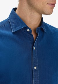 Camicia in denim blu con bottoni, dotata di un colletto classico, caratterizzata da una sottile texture e bottoni bianchi a contrasto lungo la pattina.