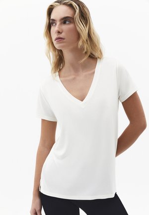 Donna bionda che indossa una maglietta bianca semplice a maniche corte con scollo a V, in piedi con le mani dietro la schiena, che guarda verso sinistra su sfondo bianco.