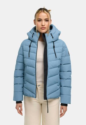 Frau trägt eine hellblaue gesteppte Jacke mit Kapuze, beige Hose und einen cremefarbenen Pullover, steht vor einem schlichten grauen Hintergrund.