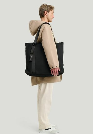 Mies, jolla on beige takki ja valkoiset housut, kantaa suurta mustaa olkalaukkua, seisoo vaaleanharmaan taustan edessä.