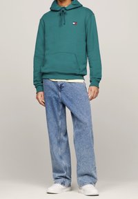 Sudadera verde con bolsillo frontal y parche de logo, combinada con vaqueros de corte holgado en azul claro y zapatillas blancas. Textura de algodón suave.