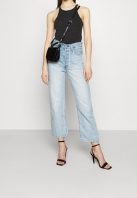 Frau trägt ein schwarzes ärmelloses Top, hellblaue hochtaillierte Jeans, schwarze Riemchenpumps und eine kleine schwarze Umhängetasche.