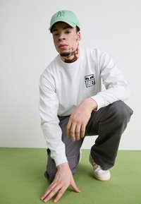 Long-sleeve white shirt with "OBEY" logo, gray pants, and a light green baseball cap. Kneeling on a green carpeted surface.