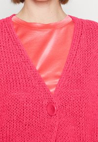 Jasně růžový pletený kardigan s výstřihem do V, se zapínáním na jediný růžový knoflík. Nošený přes lesklý růžový top, který zdůrazňuje hladkou texturu.