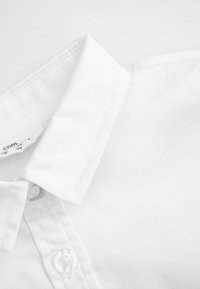 Biała bawełniana kołnierzykowa koszula z detalami w postaci guzików i przeszyć. Gładka tekstura, klasyczny design. Zbliżenie ukazujące materiał i zapięcia.