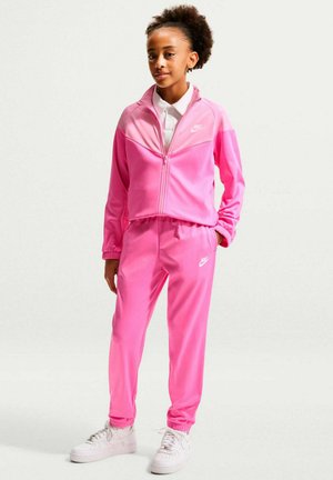 Enfant debout portant un survêtement Nike rose vif avec des baskets blanches, mains détendues, sur un fond clair uni.